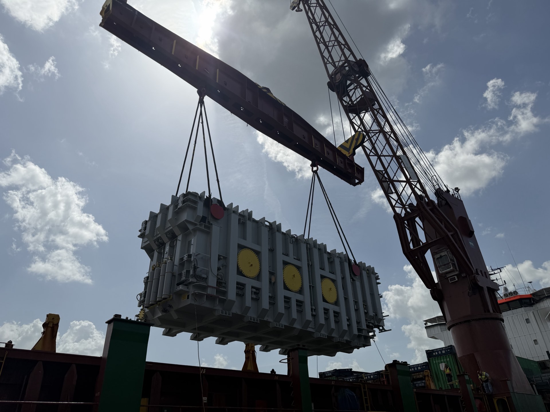 A large grey finned unit with yellow circular plates suspended from a ship’s crane on a long spreader beam, with shipping containers on deck below and a bright, partly cloudy sky.