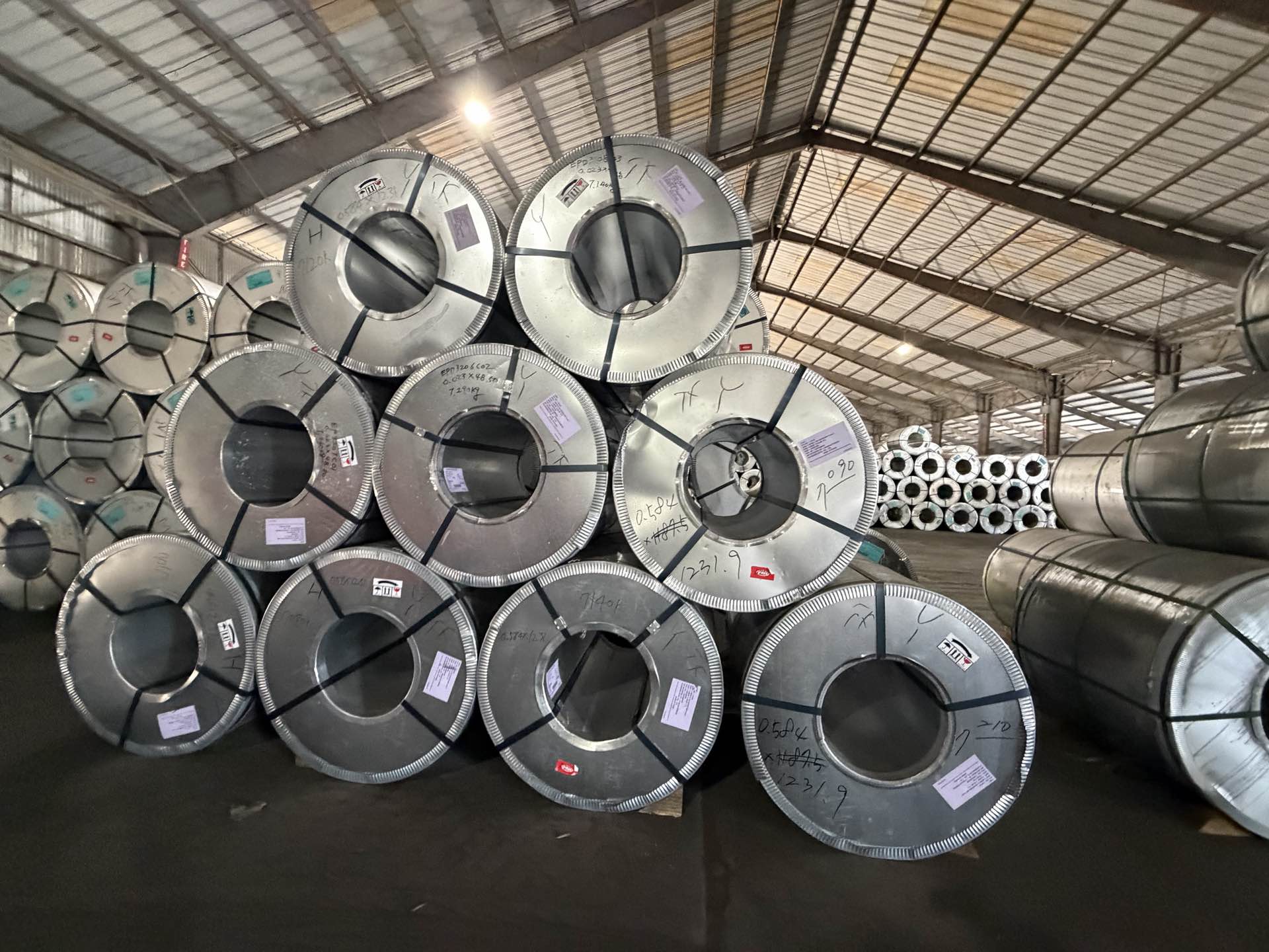 Galvanized steel coils with steel strapping and paper labels, stacked in a pyramid inside a covered storage shed.