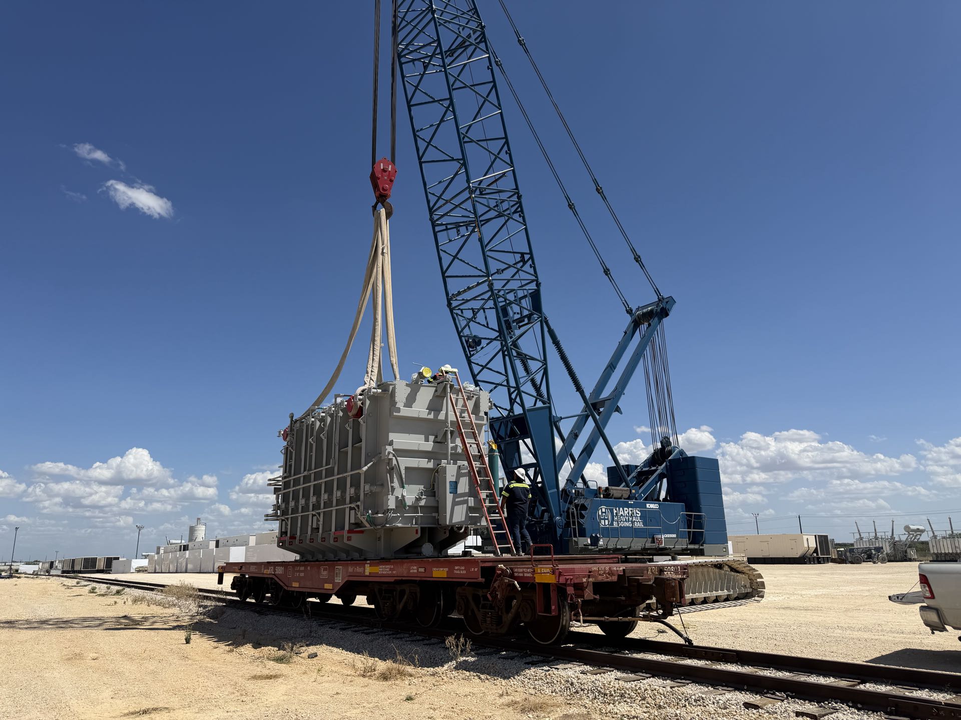 A crawler crane with a lattice boom lifting a large grey unit over a rail flatcar under a clear blue sky, with a person in a hard hat on a ladder beside the load.