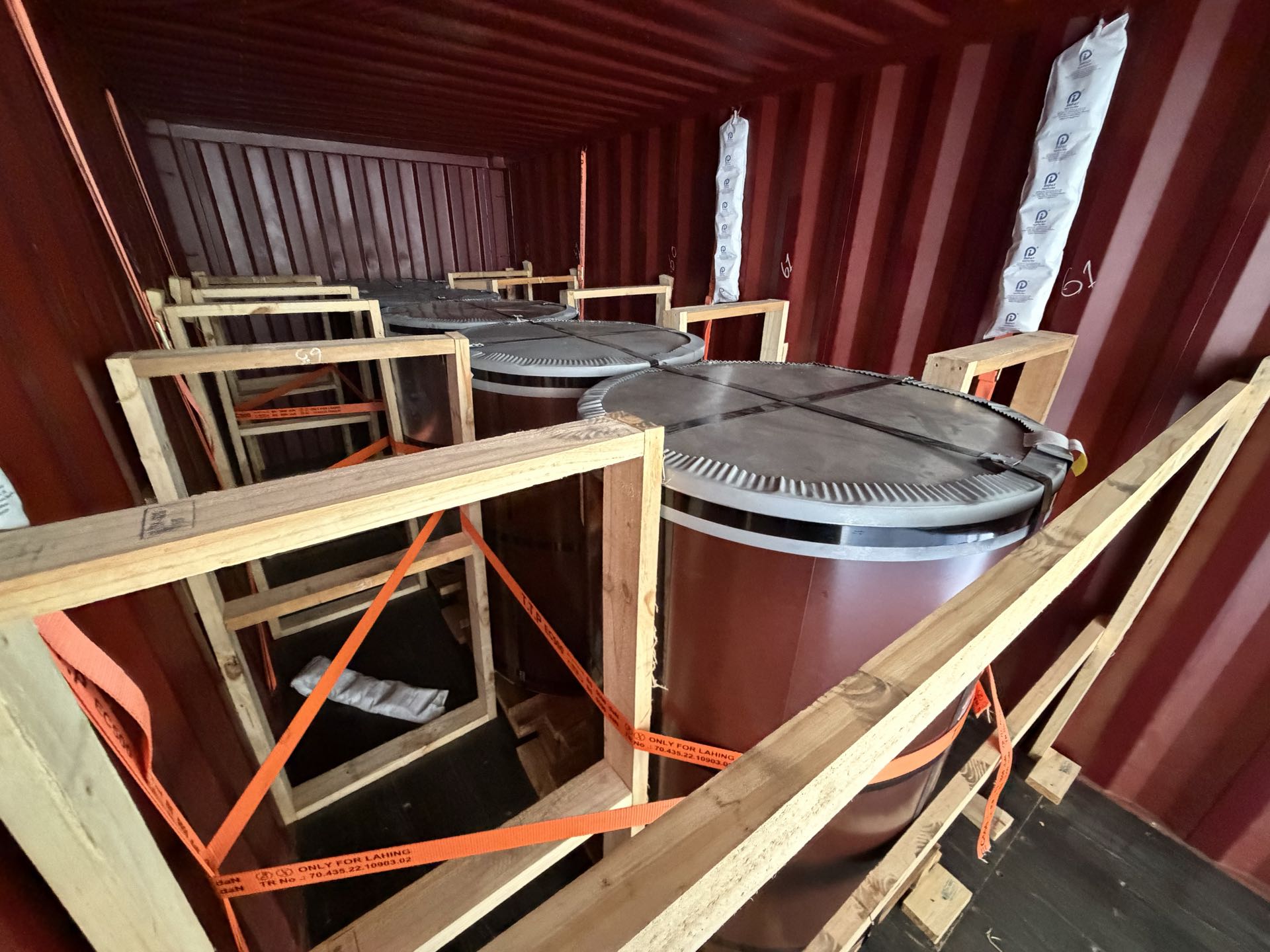 Wrapped cylindrical units standing on end inside a shipping container, blocked with timber frames and secured with orange lashing straps.