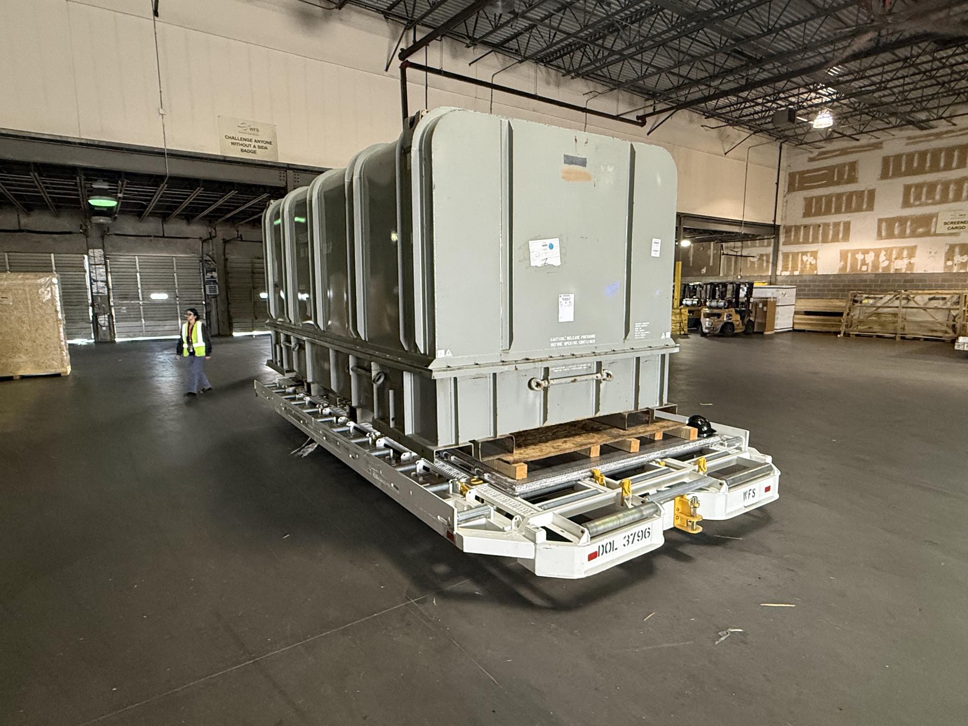 Large grey rounded-top enclosed units sitting on a white roller dolly on the floor of a cargo warehouse, with a person in a high-visibility vest walking nearby and a forklift in the background.