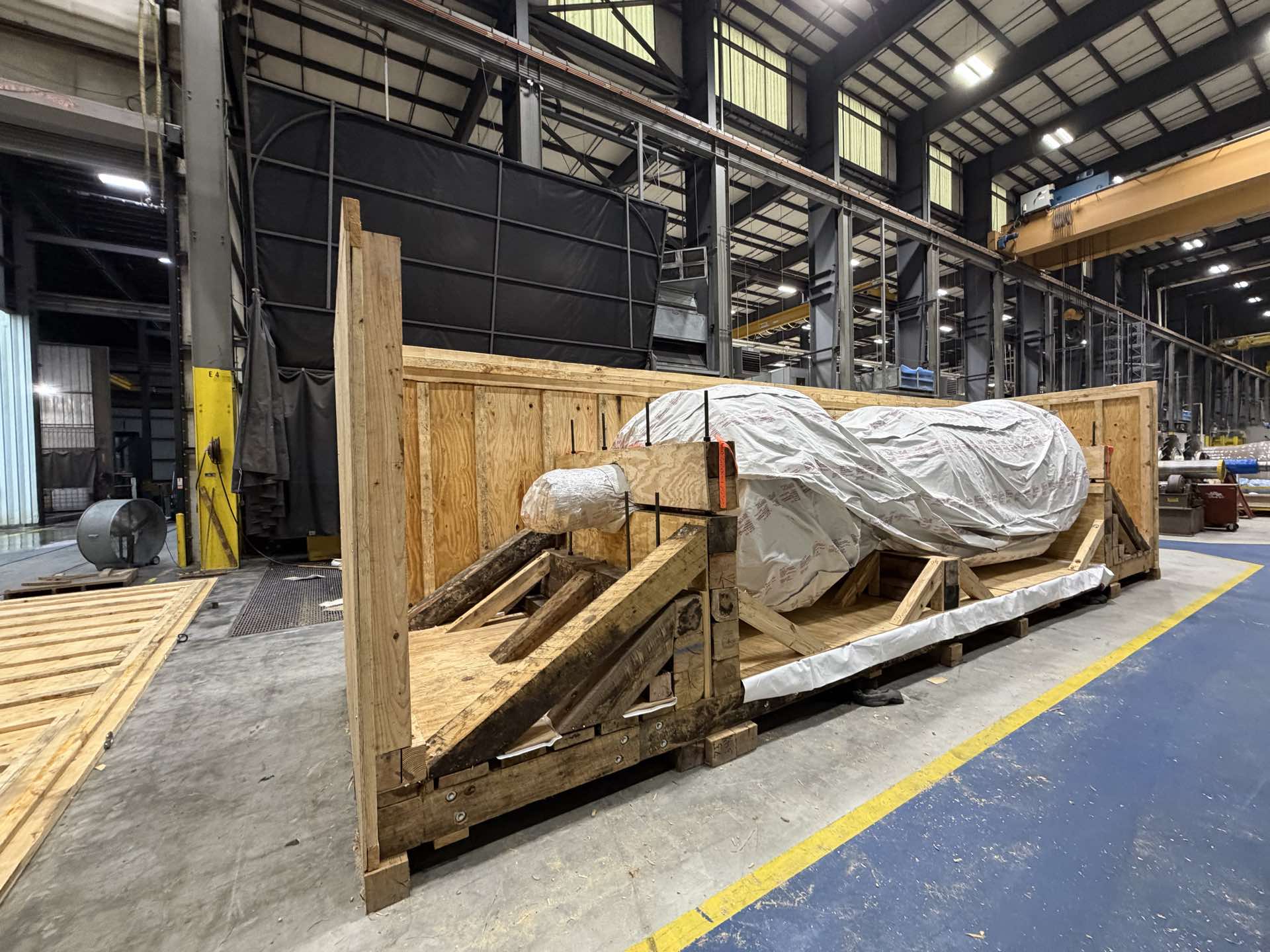 A machine wrapped in white protective film resting on timber cradles inside an open-topped wooden crate, on the floor of an industrial hall.
